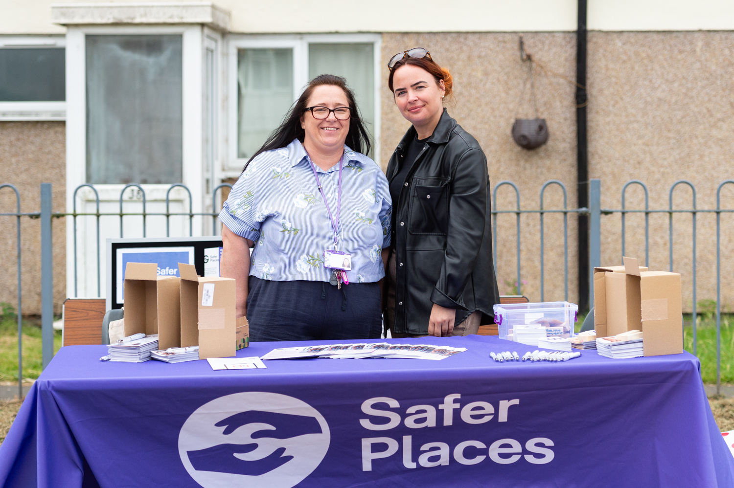 Two women smiling behind a table with a Safer Places logo. The table has four small cardboard boxes with leaflets and pens
