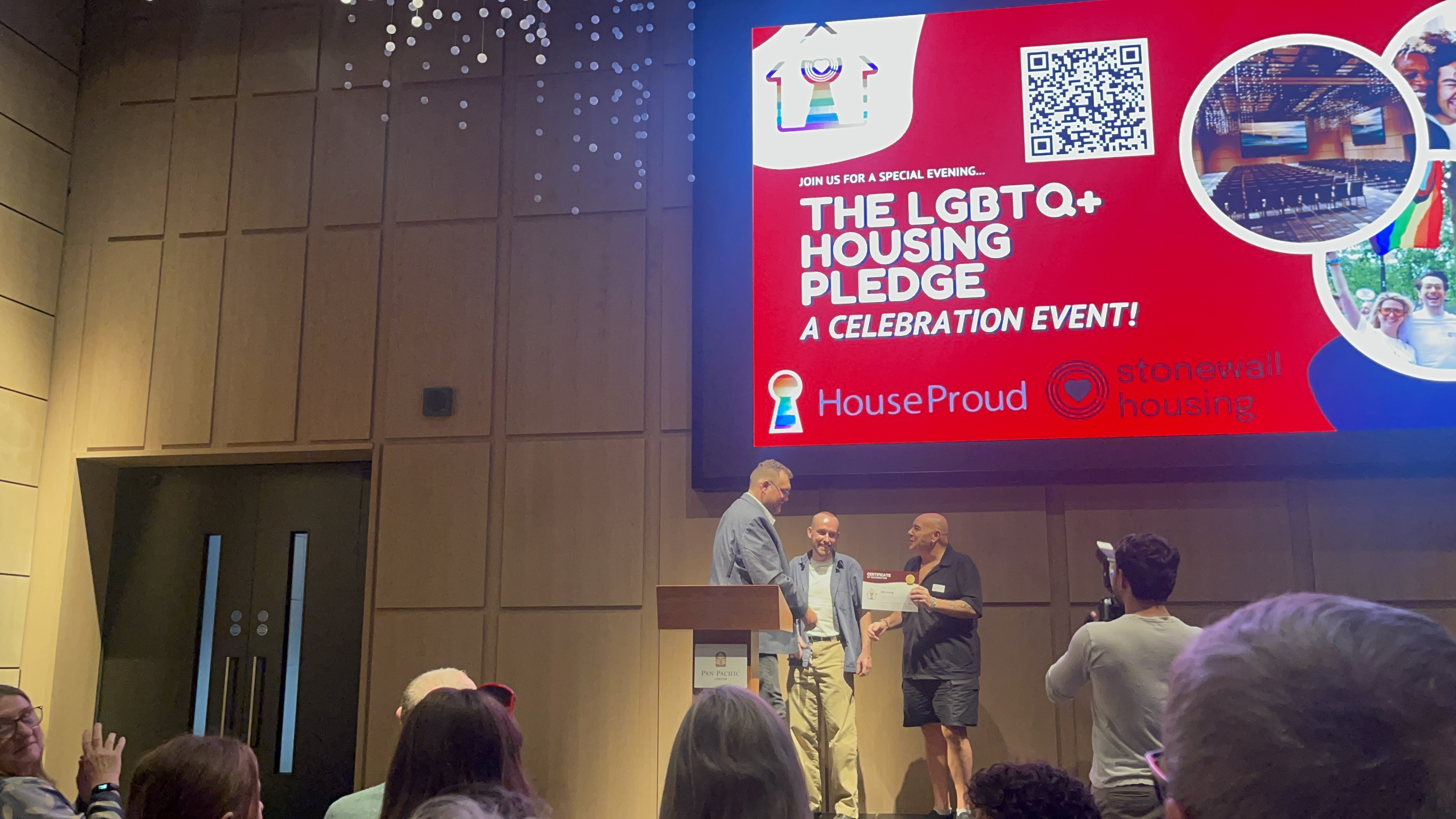 One man receiving a certificate from two men on a stage. Behind them there is a large screen reading "The LGBTQ+ Housing Pledge"
