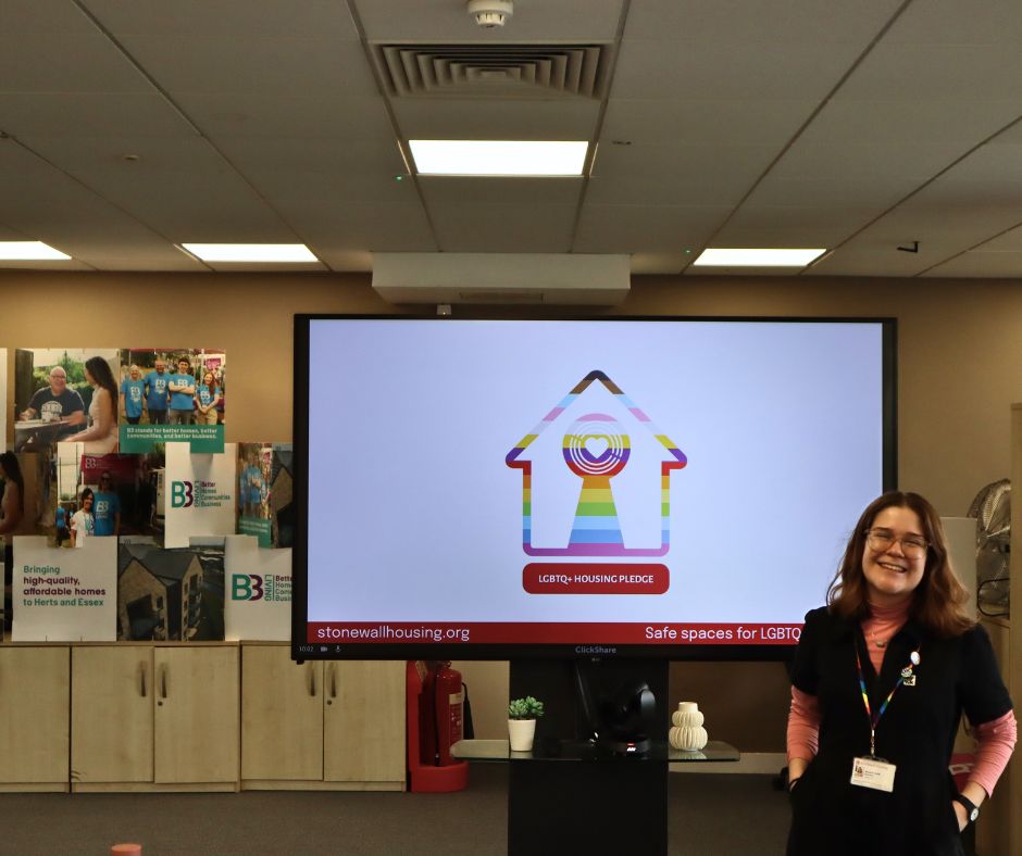 A person smiling standing to the right of a TV showing the LGBTQ+ Housing Pledge logo