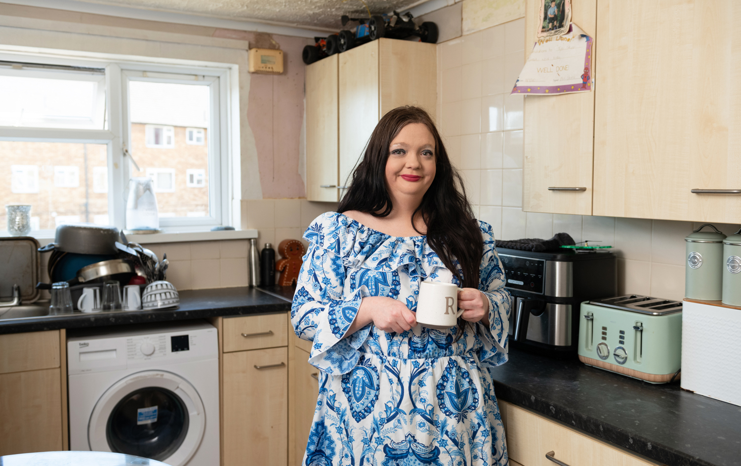 A woman wearing a blue pattern dress holding a tea mug in a kitchen