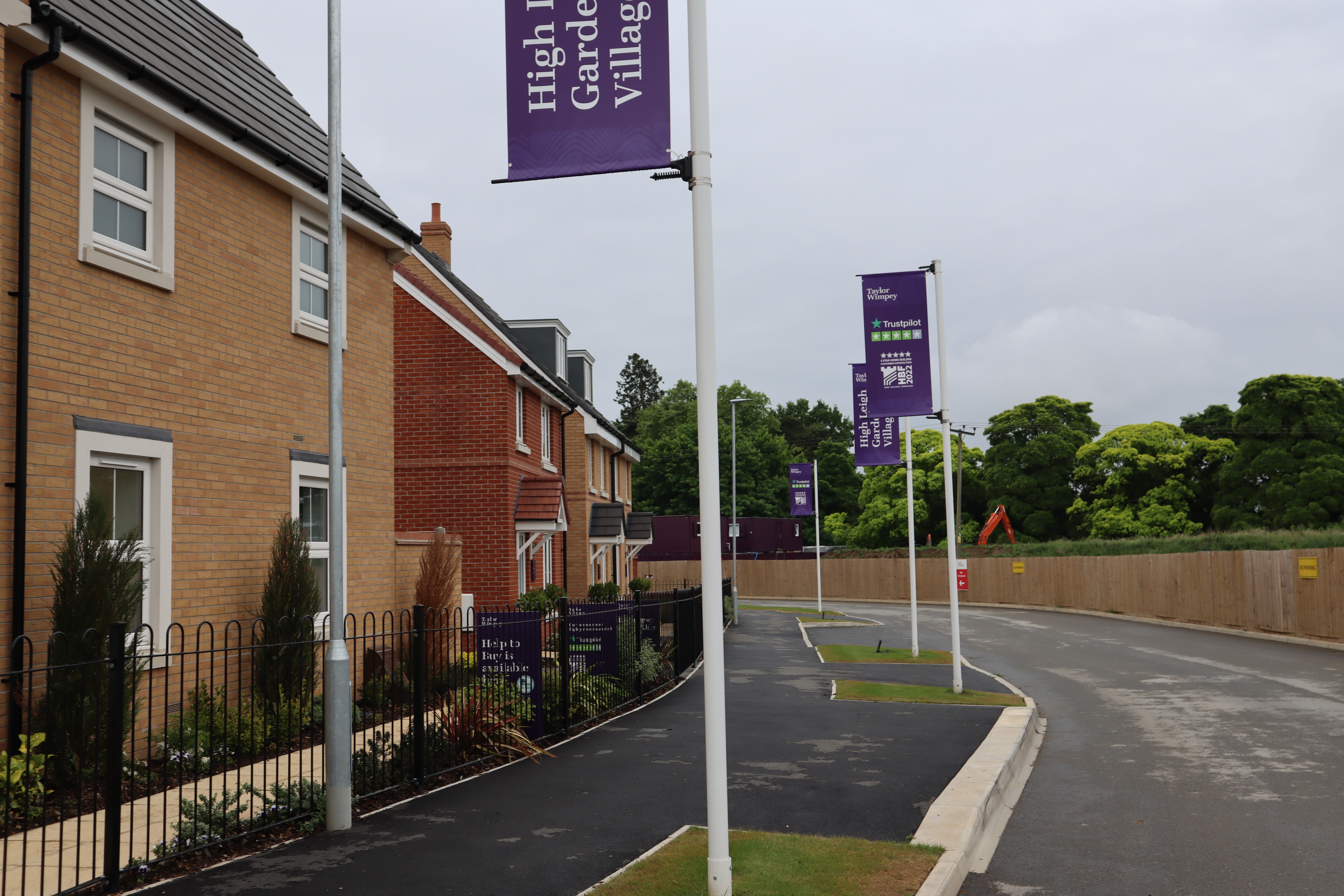 Row of semi detached houses on the left hand side of a road. There are flags on poles on the pavement that read High Leigh Garden Village. In the background, there are many trees.
