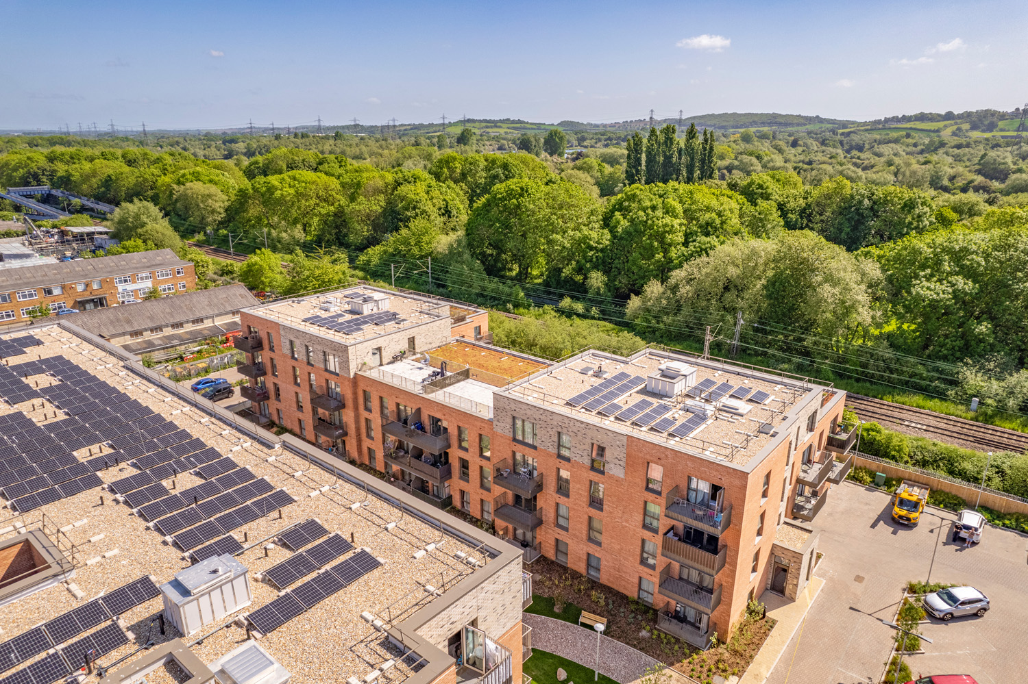 Two blocks of flats with solar panels on top. Lots of trees and a clear sky behind them