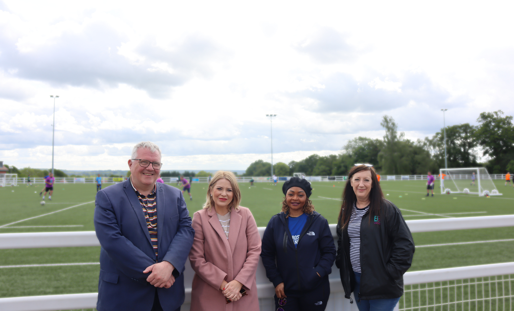Cllr Paul Seeby, Cllr Corina Gander, Tina Davies OBE, and B3Living's Community Development Manager, Helen Wright standing in front of a football pitch. Players can be seen behind them in the background